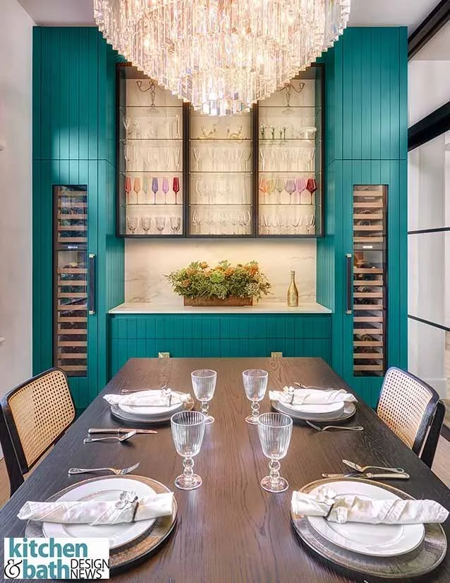 Dining table set with four place settings, clear glassware, and white napkins with cutlery, in front of a teal cabinets with a display cabinet with glass shelves and colored glassware, illuminated by a chandelier.