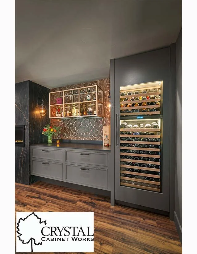 A modern kitchen corner with a built-in wine cooler, gray cabinetry, a decorative backsplash, a floating shelf with bottles and decorations, and a dark wall with a textured design.