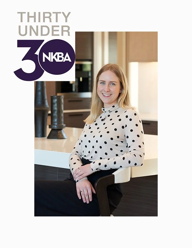 A woman with blonde hair wearing a white blouse with black polka dots, smiling, sitting in a modern kitchen with dark wood cabinets and decorative vases, celebrating NKBA's 30 Under 30.