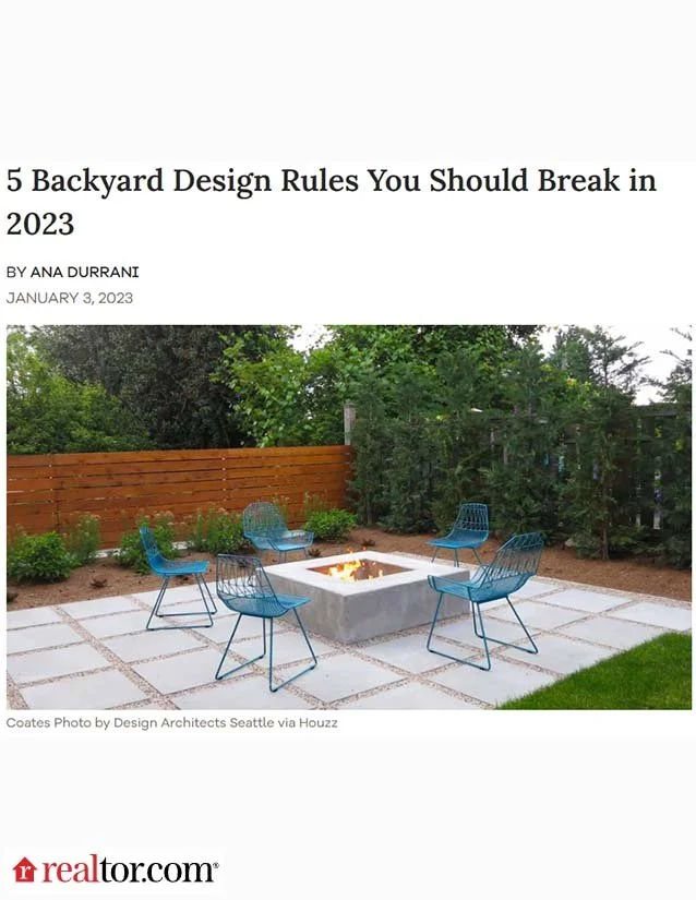 A backyard patio with six blue wire chairs arranged around a square fire pit, surrounded by a mix of grass, paving stones, and a wooden fence with green bushes and trees in the background.