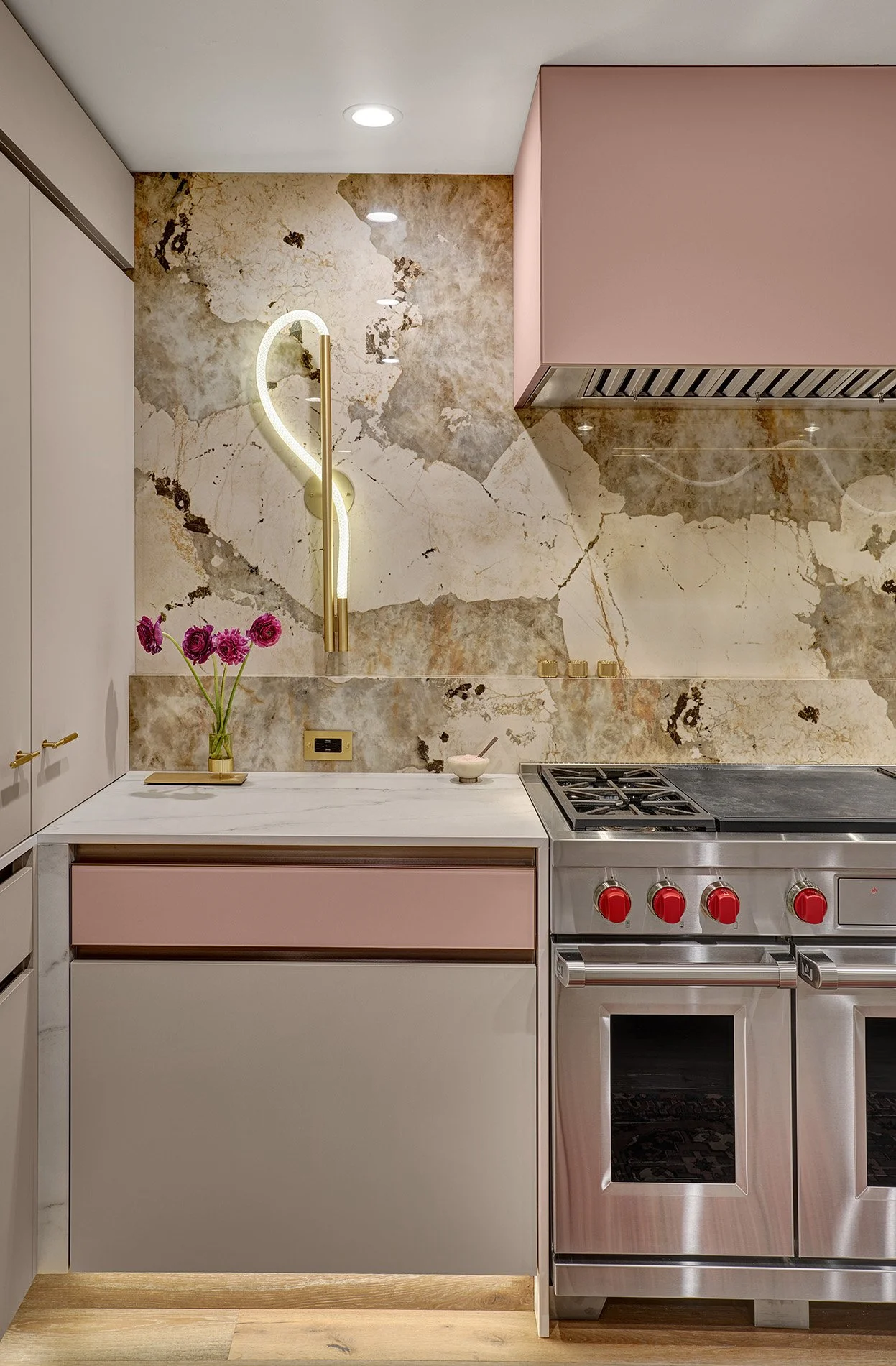Modern kitchen with pink and white cabinetry, a marble countertop, a Dekton backsplash, and a stainless steel oven with red knobs. There is a vase of pink flowers and a decorative wall light.