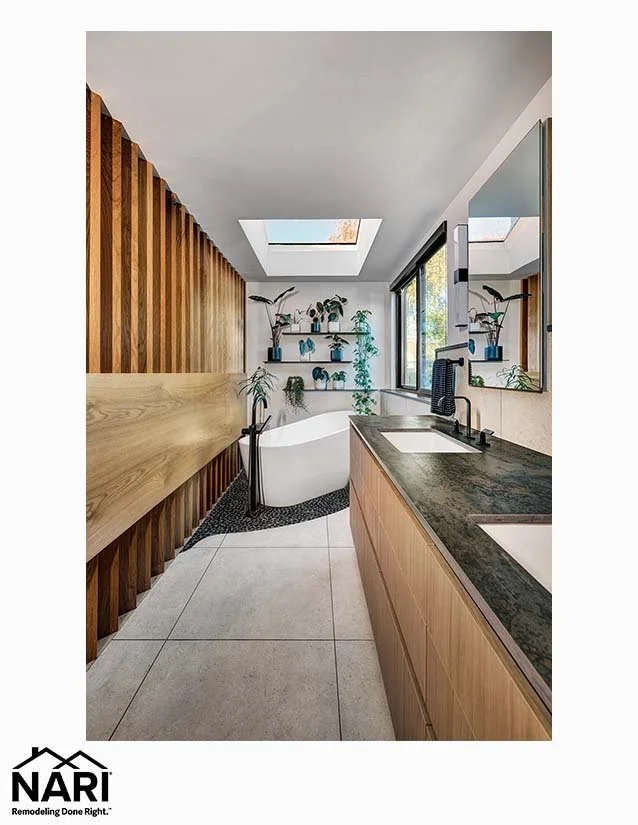 Modern bathroom with white freestanding bathtub near a window, wooden wall paneling, beige tile flooring, black countertop with sink, large mirror, and floating shelves with plants and decorative items.