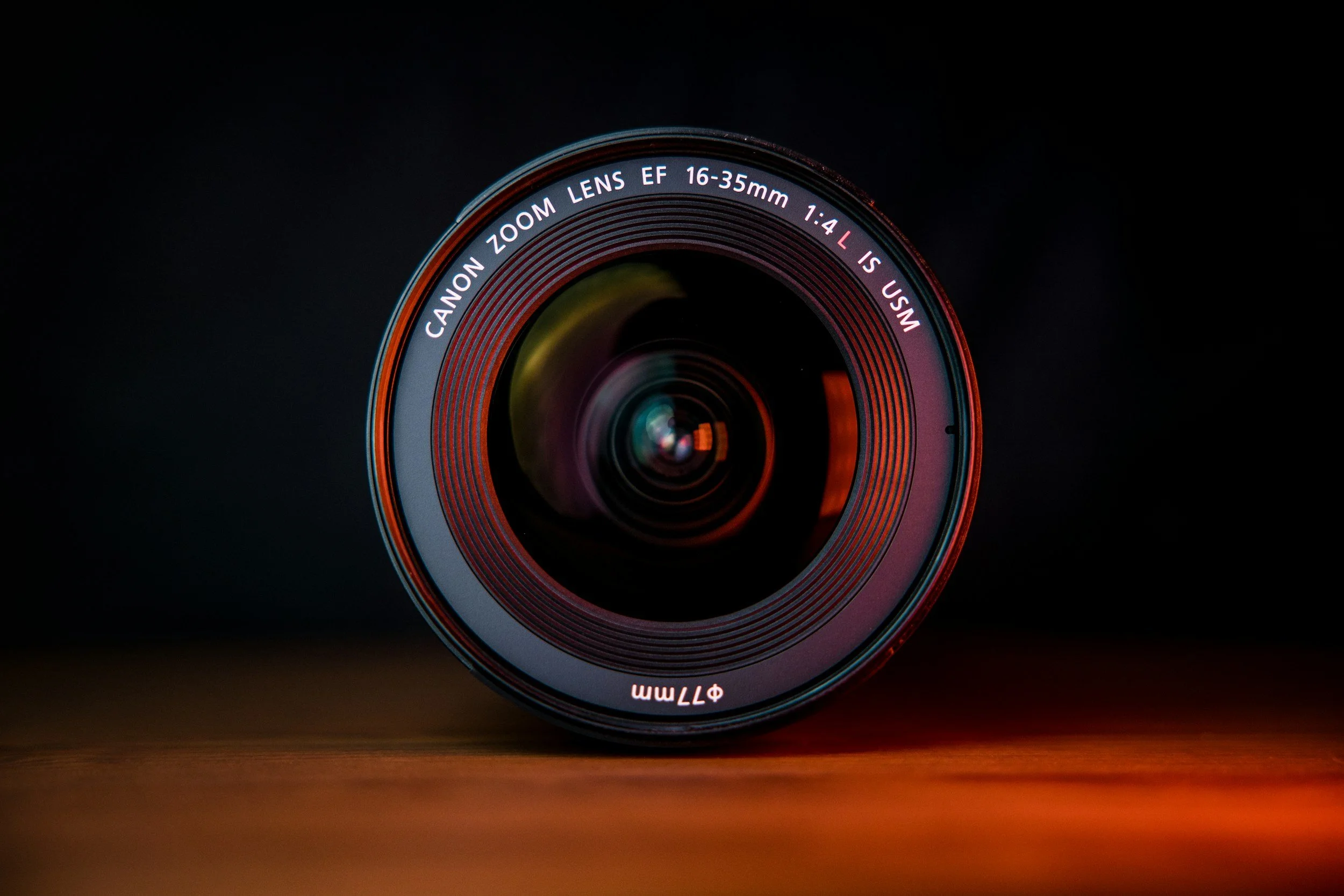 Close-up of a Canon zoom lens with 16-35mm focal length, on a wooden surface against a dark background.