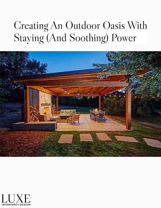 An outdoor covered patio with seating and dining furniture, surrounded by a lawn and trees at dusk.